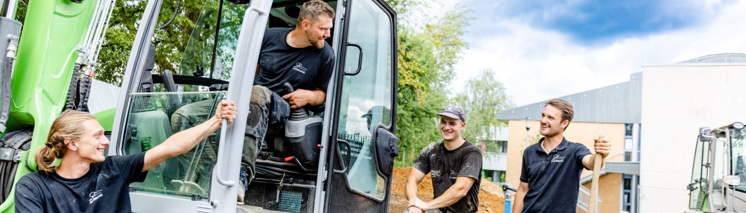Steinhake-Team auf der Baustelle. Einer sitzt im Bagger, drei andere unterhalten sich mit ihm.